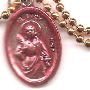 May include: A pink oval-shaped pendant with a gold chain. The pendant features a depiction of a saint with a halo and a book. The text "St. Lucy" is inscribed on the pendant.