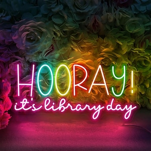 Puede incluir: Un letrero de neón vibrante con la palabra "HOORAY!" en colores brillantes, con el texto "it's library day" debajo. El letrero está sobre un fondo de rosas coloridas, con un degradado de rosa, amarillo y verde.