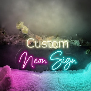 May include: Custom Neon Sign with the words "Custom Neon Sign" in white neon lettering on a clear acrylic background. The sign is illuminated with pink and blue neon lights.