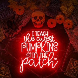 May include: A red neon sign with the text "I teach the cutest pumpkins in the patch" in white script. The sign is surrounded by Halloween decorations, including pumpkins, a skull, and tombstones.