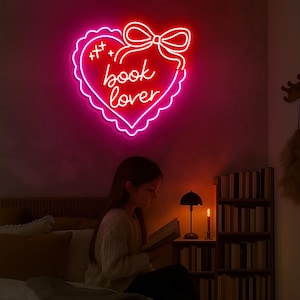 May include: A neon sign in the shape of a heart with the words "book lover" in cursive script. The sign is outlined in pink and red, with a bow at the top. A person is reading a book under the sign.