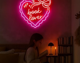May include: A neon sign in the shape of a heart with the words "book lover" in cursive script. The sign is outlined in pink and red, with a bow at the top. A person is reading a book under the sign.