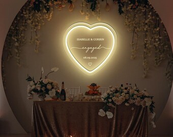 May include: A heart-shaped neon sign with the word "engaged" and names "Isabelle & Corbin" above a table set for a celebration. The table is covered with a gold sequined cloth and surrounded by flowers and candles.
