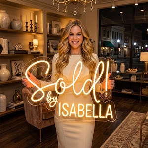 Pode incluir: Uma placa de néon com as palavras "Sold by ISABELLA" em itálico. A pessoa segura a placa em frente a si. A placa emite uma luz amarela quente. O fundo mostra uma sala com prateleiras e uma janela.