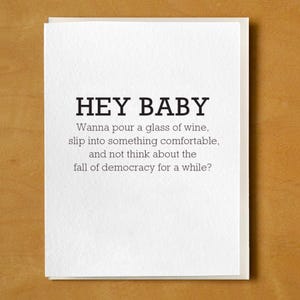May include: A white greeting card with the text "HEY BABY" in bold, black letters. Below, the card reads: "Wanna pour a glass of wine, slip into something comfortable, and not think about the fall of democracy for a while?"