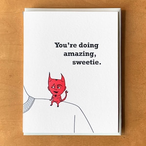 May include: A greeting card with a red devil cartoon character on a white shirt. The card reads "You're doing amazing, sweetie."