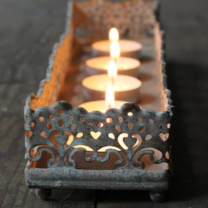 May include: A rustic metal candle holder with a decorative, lacy design. Four white tea lights are placed inside the holder, with the flames glowing brightly.