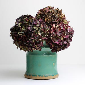 May include: A teal ceramic vase with a rustic finish holds a bouquet of dried hydrangeas with burgundy and purple flowers.