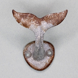 May include: A metal wall hook in the shape of a whale's tail. The tail is brown and white, with a textured, distressed finish. The base is oval with two holes for mounting.