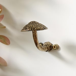 May include: A wall-mounted hook shaped like a mushroom with a textured, antique bronze-coloured finish. The hook features a wide, domed cap and a curved stem that attaches to the wall with a small circular base secured by two visible metal screws.