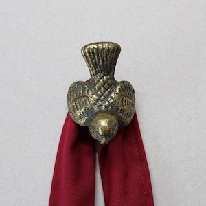 May include: A brass bird ornament with detailed wings and a textured finish, attached to a deep red fabric. The bird is a decorative item. The fabric is folded and hangs down from the bird.