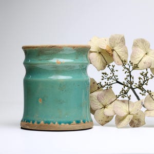 May include: A teal ceramic vase with a textured rim and a slightly distressed finish. The vase is sitting on a white surface next to a sprig of dried white flowers.