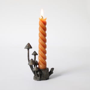 May include: An orange, twisted candle is lit, set in a bronze-coloured mushroom-themed candle holder. The candle holder is shaped like a tree stump with several mushroom accents. The candle is approximately 20 cm tall.
