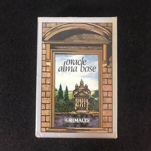 May include: A box for the "Oracle Alma Bosa" tarot deck. The box is white with a silver border and features a picture of a building with a window. The window shows a view of a building with a dome and a river. The text "Grimaud" is printed on the box.