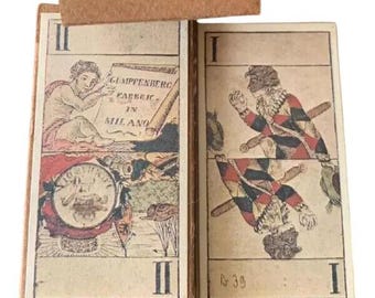 May include: Antique playing cards with detailed illustrations. One card features a figure with a scroll, the text "GUMPFFENBERG FABBRIC IN MILANO", and a circular emblem. The other card shows a harlequin figure. The cards are in a worn, vintage condition.