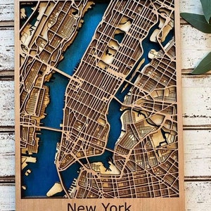 Puede incluir: Mapa de Nueva York cortado con láser en madera, con detalles intrincados de calles y edificios. El mapa tiene un marco de madera natural y un fondo azul que representa el agua. El texto "New York" y las coordenadas están en la parte inferior.