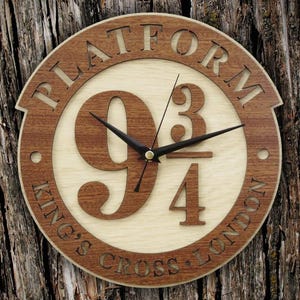 May include: Wooden wall clock with a circular design. The clock features the words "PLATFORM" and "KING'S CROSS LONDON" in a dark brown, laser-cut font. The clock face displays the numbers "9 3/4" in a lighter wood tone, with black clock hands.