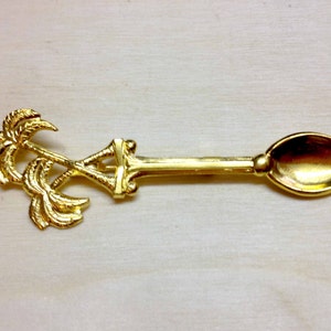 Könnte beinhalten: Ein goldfarbener Löffel mit Palmenmuster am Griff. Der Löffel hat einen langen, schlanken Griff und eine kleine, oval geformte Schüssel. Das Palmenmuster ist detailliert und verleiht dem Löffel ein dekoratives Element.