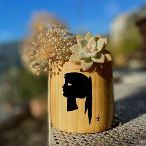 Può includere: Un vaso cilindrico in legno chiaro con una silhouette nera del profilo di una donna. Il vaso contiene una succulenta e fiori secchi. Lo sfondo è sfocato con un cielo blu.