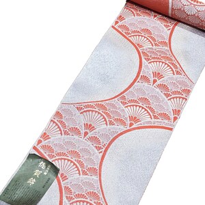May include: Rolled-up Japanese obi with a silver background and a repeating fan pattern in coral and white. The obi has a coral-colored edge and a small tag with Japanese text.