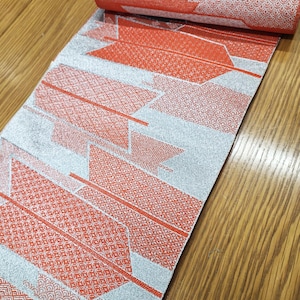 Op de afbeelding: Opgerolde decoratieve stof met een herhalend patroon van rode en zilveren geometrische vormen. De stof is gedeeltelijk uitgerold en onthult het ingewikkelde ontwerp. Het kleurenpalet omvat tinten rood en zilver, waardoor een visueel aantrekkelijk textiel ontstaat.