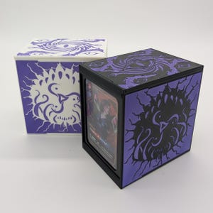 Puede incluir: Dos cajas cúbicas para guardar cartas. Una es blanca con diseños morados, la otra es negra y morada. Ambas cajas presentan un diseño de ojo en la tapa y un diseño estilizado de pájaro en los laterales. Una carta es visible dentro de la caja negra.