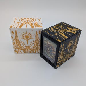 Puede incluir: Dos cajas rectangulares para guardar cartas, una blanca con líneas doradas y otra negra con líneas doradas. Ambas presentan un motivo de sol y espada. La caja negra tiene una ventana frontal transparente que muestra una carta coleccionable.