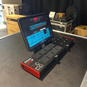 May include: A black and red Akai LPD8 pad controller with a tablet attached to the top. The tablet is displaying a music production app with a blue and white interface.