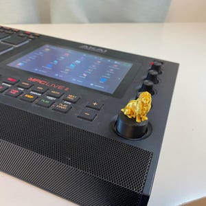 May include: Black MPC Live II music production center with a gold-colored lion figurine on a knob. The device has a screen displaying a user interface and various buttons labeled with functions. The lion figurine adds a decorative touch to the instrument.