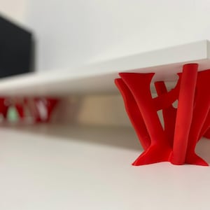 Puede incluir: Una estantería blanca con patas abstractas rojas impresas en 3D. La estantería está sostenida por cinco patas, cada una con un diseño único.