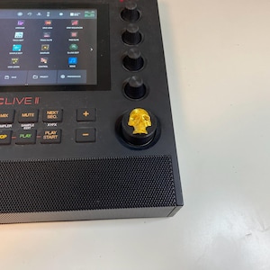 May include: A black MPC Live II music production sampler with a gold skull knob. The screen displays the menu options for the device.