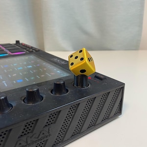 Peut inclure: Un contrôleur de production musicale noir avec un dé doré sur l'un des boutons. Le contrôleur a un écran qui affiche une grille de carrés. Le texte "MPC Live" est visible sur le contrôleur.