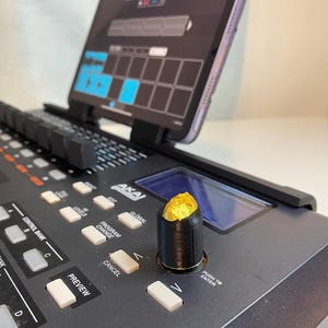 Peut inclure: Un clavier de production musicale noir avec une tablette fixée en haut. Le clavier possède une variété de boutons et de molettes, dont une grosse molette avec un dessus doré.