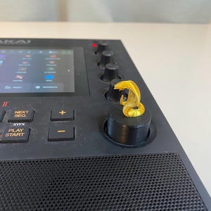 May include: Akai Live II music production workstation with a gold knob with a snake design on top. The workstation has a black and gray color scheme and features a touchscreen display, buttons, and knobs.