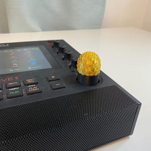 May include: A black Akai MPC Live II music production sampler with a gold knob on the top. The screen displays a grid of icons. The buttons below the screen read "Sampler", "Mute", "Play", and "Start".