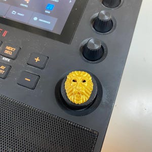 May include: Close-up of a black electronic music production device with a gold lion head knob. The device has buttons labeled "MUTE", "NEXT SEQ.", and "PLAY START". The screen displays "MPC LIVE II".