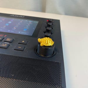 May include: A black music production controller with a gold turtle knob. The controller has a screen that displays "MPC LIVE II" and buttons labeled "Sample", "Play", and "Start".