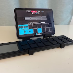 May include: A black Korg NanoPAD2 MIDI controller with a tablet displaying a music production interface. The tablet screen shows a grid of pads and waveform visualizations in blue and white. The setup is on a white surface.