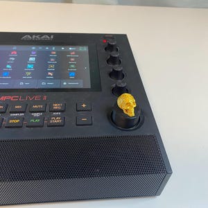 May include: A black Akai MPC Live II music production sampler with a gold skull knob. The screen displays a grid of icons and the text "MPC Live II".