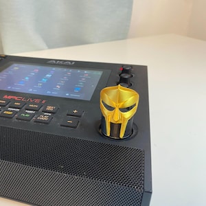 May include: A gold-colored 3D printed mask knob for a music production controller. The mask is a stylized representation of a warrior's helmet. The knob is attached to a black music production controller with the text "MPCLIVE II" on the front.