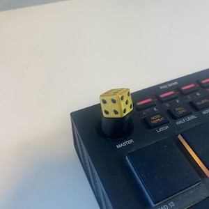 Puede incluir: Un controlador de producción musical negro con una perilla amarilla en forma de dado. La perilla está etiquetada como "Master". El controlador tiene botones etiquetados como "Note Repeat", "Latch", "Half Level" y "Notes".
