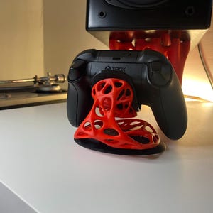 May include: A black Xbox controller is resting on a red, 3D printed stand with a lattice design. The stand has the word "Xbox" printed on it.
