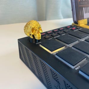 Peut inclure: Un bouton en forme de crâne doré est placé sur un pad de production musicale noir avec une variété de boutons et de pads. Le pad est éclairé d'une lumière jaune. Le bouton en forme de crâne est un accessoire décoratif pour le pad de production musicale.