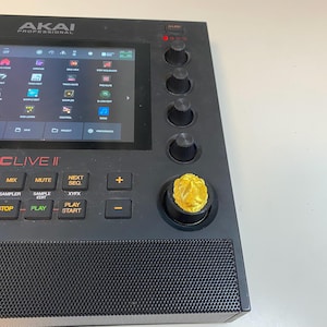 May include: Black Akai MPC Live II music production sampler with a gold knob. The screen displays a menu with options for track editing, sample editing, and control.