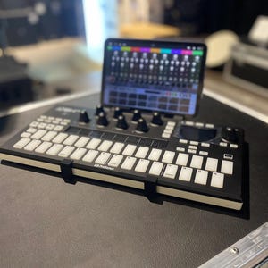 May include: A black and white electronic music keyboard with a white screen displaying a colorful grid pattern. The keyboard has a black and white design with a white border. The keyboard is sitting on a black surface.