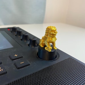 May include: A gold lion figurine sits on top of a black knob on a music production device. The device has a screen, buttons, and knobs. The words "Next Seq." and "Play Start" are visible on the device.
