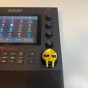May include: A black Akai MPC Live II music production sampler with a gold 3D printed mask knob. The screen displays the menu options for the device. The buttons on the device are labeled: Menu, Main, Mix, Mute, Next Seq, Browse, Grid, Sampler, Sample Edit, X/YFX, Rec, Over Dub, Stop, Play, Play Start.