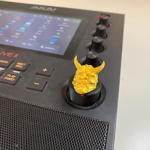 May include: A gold-colored 3D printed knob with a detailed demon face design, mounted on a black knob. The knob is on a black music production device with a screen displaying the text "MPCLIVE II" and "AKAI".