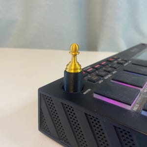 Peut inclure: Un bouton en forme de pièce d'échecs dorée sur un contrôleur de production musicale noir. Le contrôleur possède une grille de boutons avec des lumières qui brillent.
