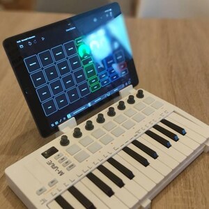 May include: A white MIDI keyboard with 25 keys and a grid of buttons. A tablet is attached to the keyboard and displays a music production app.